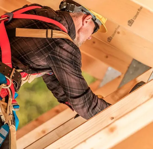 Construction worker wearing safety gear installs wooden beams on a roof structure outdoors.