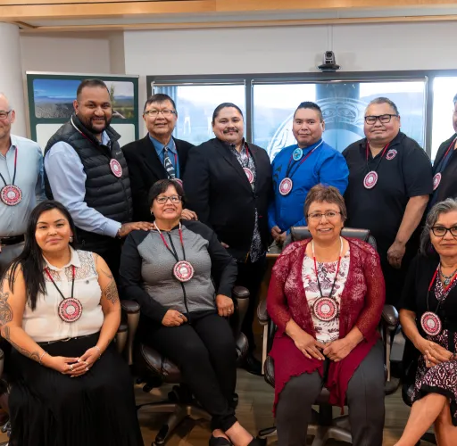 A group of thirteen adults pose together in an office setting, each wearing a lanyard with a circular badge. Some are seated while others stand behind them.