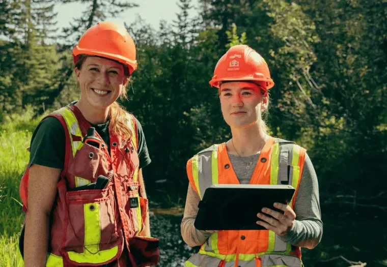 Two individuals wearing orange safety gear and helmets stand outdoors. One is holding a tablet. Trees and greenery are visible in the background.