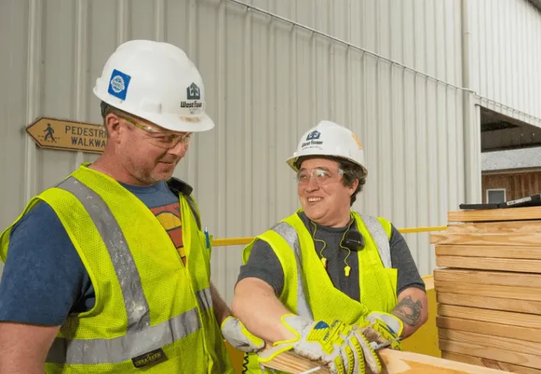 Two construction workers in safety vests, gloves, and helmets are handling wooden planks outside a building. One worker is smiling and talking to the other. A "Pedestrian Walkway" sign is visible.