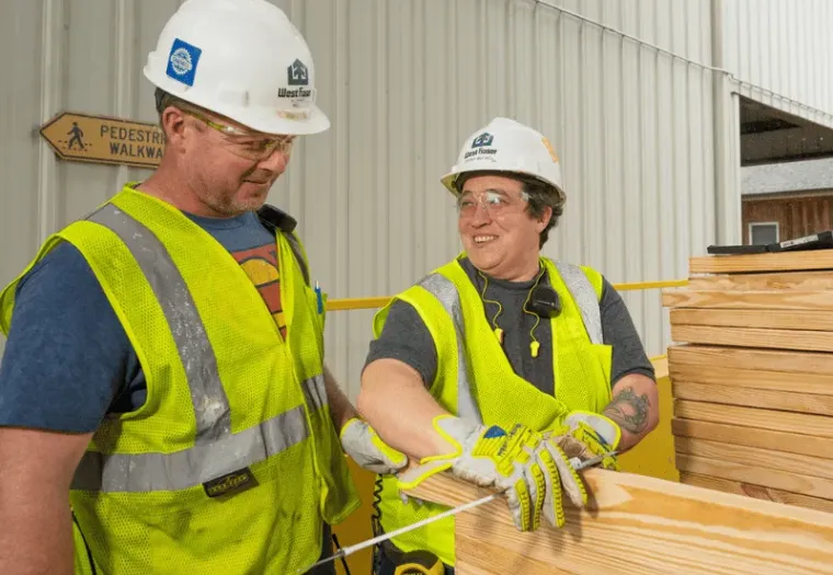 Two construction workers in safety vests, gloves, and helmets are handling wooden planks outside a building. One worker is smiling and talking to the other. A "Pedestrian Walkway" sign is visible.
