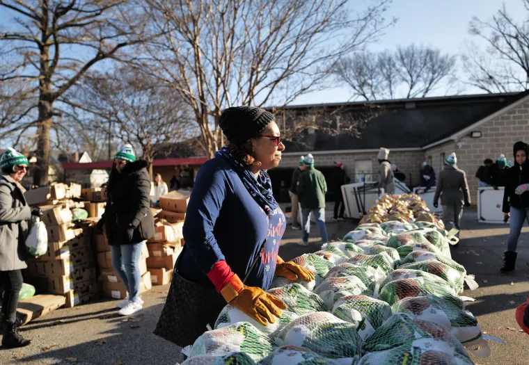 A person stands near a table filled with packaged food items outdoors. Other individuals are visible in the background, also participating in the activity on a clear day.