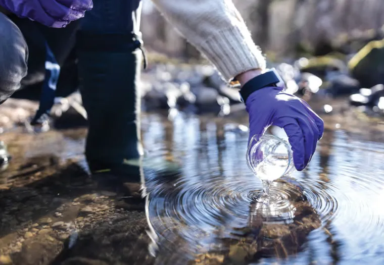 A person wearing purple gloves and a white sweater kneels by a rocky stream, collecting a water sample in a clear glass jar.