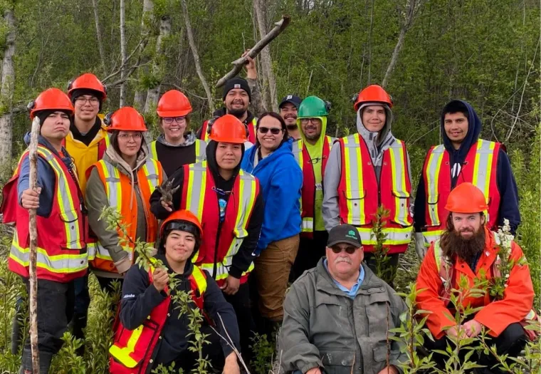 A group of people wearing orange safety vests and hard hats stand outdoors in a forested area, posing for a photo. Some are holding tools, surrounded by trees and vegetation.