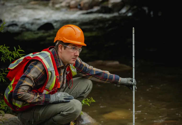 A West Fraser worker wearing a safety vest, helmet, and gloves kneels by a stream, measuring the water level with a white and black striped rod.