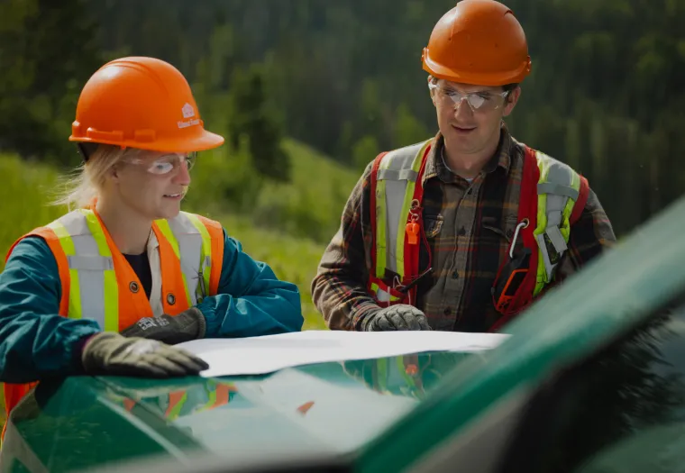 Two construction workers wearing safety helmets and high-visibility vests review plans on the hood of a green vehicle in an outdoor setting.