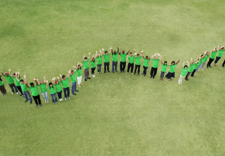 A group of people wearing green shirts stand outside on grass, forming a wavy line and raising their hands in the air.