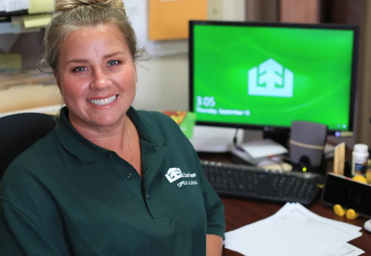 A person smiles while sitting at a desk with a computer in the background displaying a green screen. They are wearing a green polo shirt with a logo and name badge.
