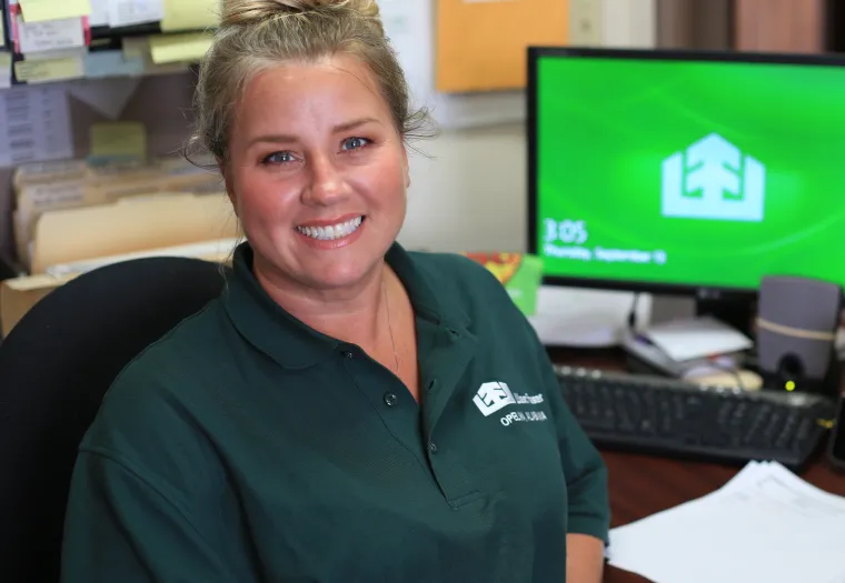 A person smiles while sitting at a desk with a computer in the background displaying a green screen. They are wearing a green polo shirt with a logo and name badge.
