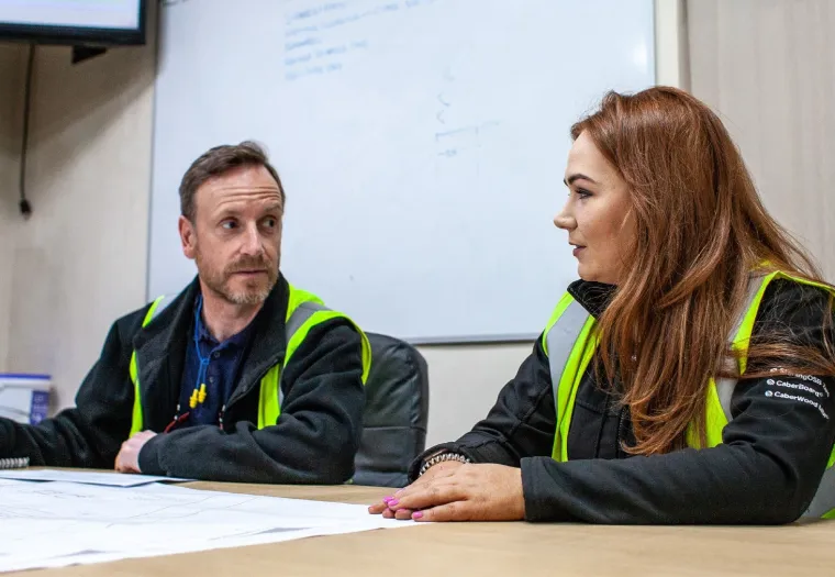 Two people wearing high-visibility vests are seated at a table, discussing documents. A whiteboard with notes is visible in the background.