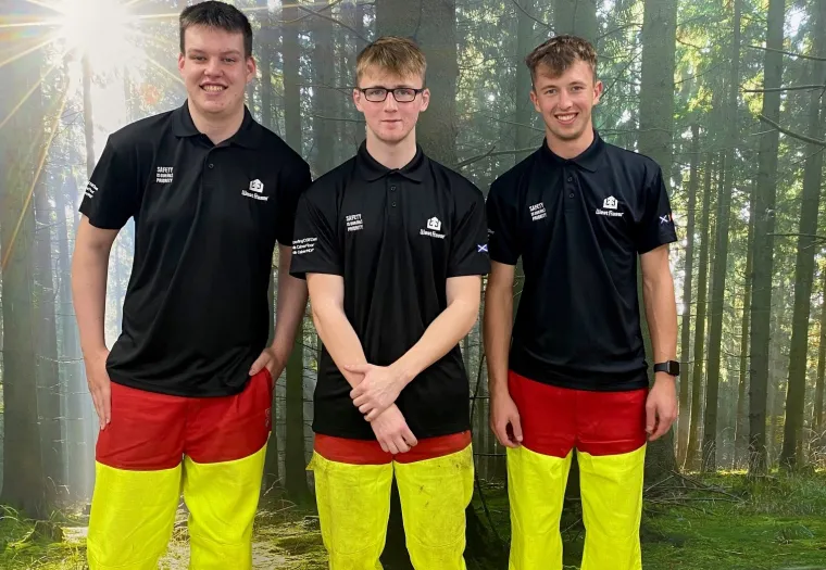 Three young people wearing black shirts and bright yellow pants with red accents stand in front of a forest-themed wall. They are positioned side by side, smiling towards the camera.