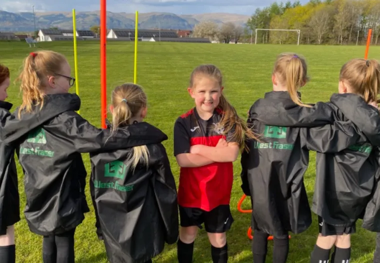 A group of children wearing sports jackets stand in a field with their arms around each other. One child in the center, wearing a different outfit, stands with arms crossed.