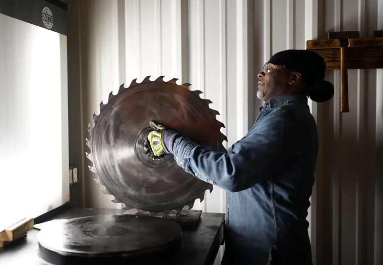 A person wearing a denim shirt and protective gloves holds a large circular saw blade inside a workshop.