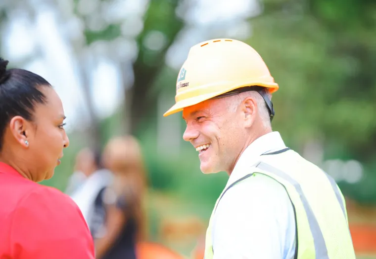A man wearing a hard hat and safety vest smiles while talking to a woman in a red jacket at an outdoor setting.