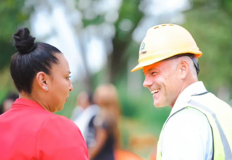A man wearing a hard hat and safety vest smiles while talking to a woman in a red jacket at an outdoor setting.