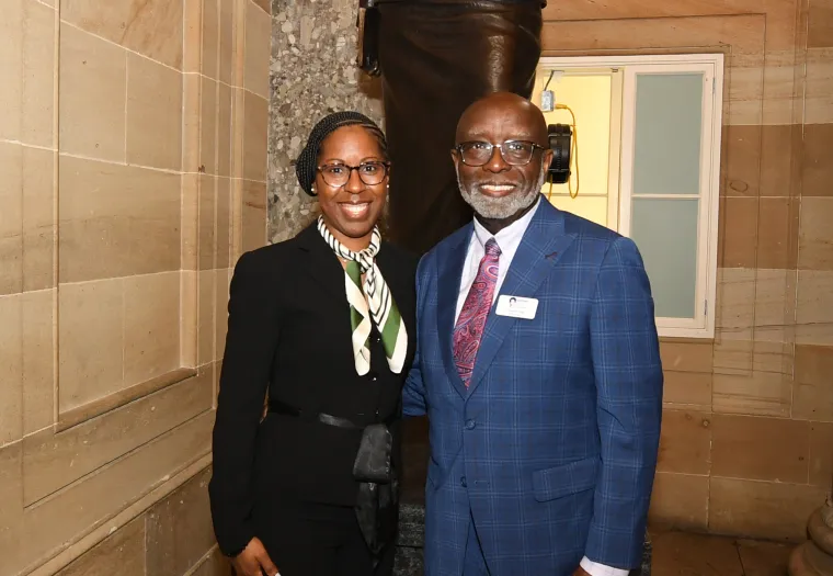 Two people stand in front of a statue in a well-lit, ornate hallway. The individual on the left is dressed in a black suit with a patterned scarf, and the one on the right is dressed in a blue suit.