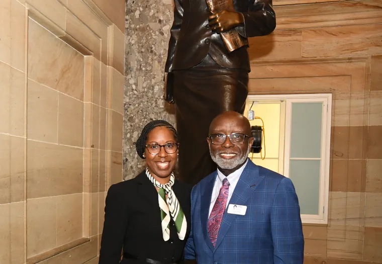 Two people stand in front of a statue in a well-lit, ornate hallway. The individual on the left is dressed in a black suit with a patterned scarf, and the one on the right is dressed in a blue suit.