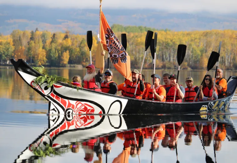 A group of people wearing life jackets sit in a decorated canoe holding paddles and an orange flag, on a calm body of water with autumn trees in the background.