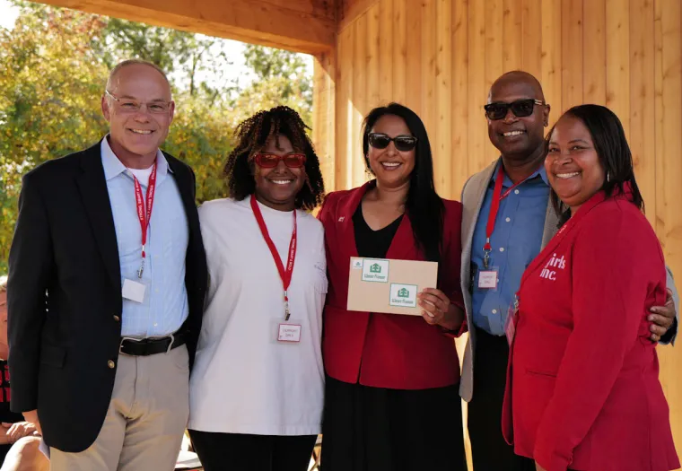 Five individuals wearing name badges stand together smiling, with one holding a certificate, in an outdoor setting with wooden walls.