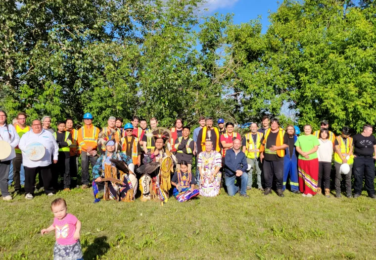 A group of people, including various adults in construction vests and traditional attire, pose outdoors in front of trees. A child in a pink top and blue skirt stands in the foreground.