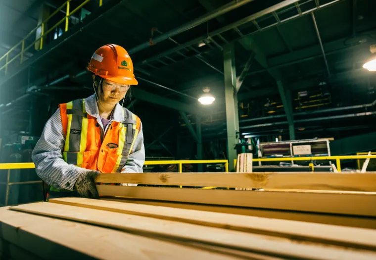 A West Fraser worker wearing a hard hat and safety vest examines wooden planks in a mill setting with machinery and pipes in the background.