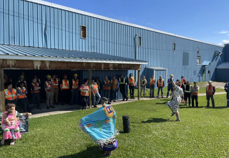 A group of people wearing safety vests stand outside a blue industrial building, observing two dancers in colorful regalia performing on a grassy area. A child sits nearby watching the performance.