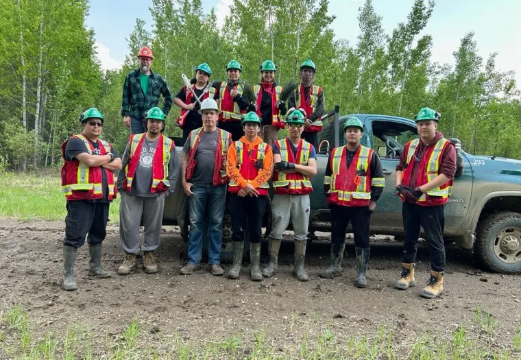 A group of eleven people in safety gear and hard hats standing together in front of a truck in a forested area.