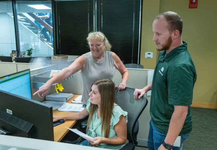 Three people are gathered around a desk in an office setting, with two standing and one seated, looking at a computer screen. One person is pointing at the monitor.