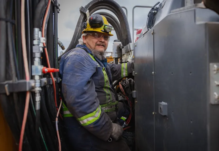 An older man wearing a hard hat and protective gear works on heavy machinery, surrounded by cables and industrial equipment.