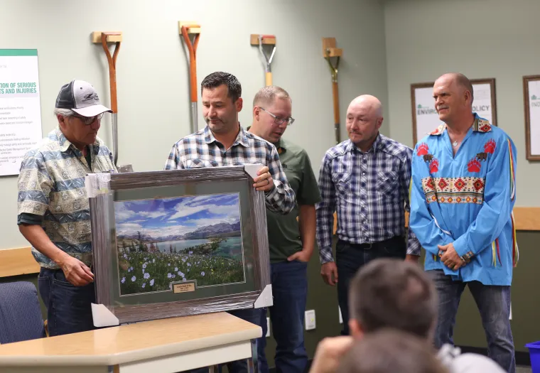 Four men stand together while one presents a framed landscape painting to another. They are in an indoor setting with a group of onlookers seated in front of them.