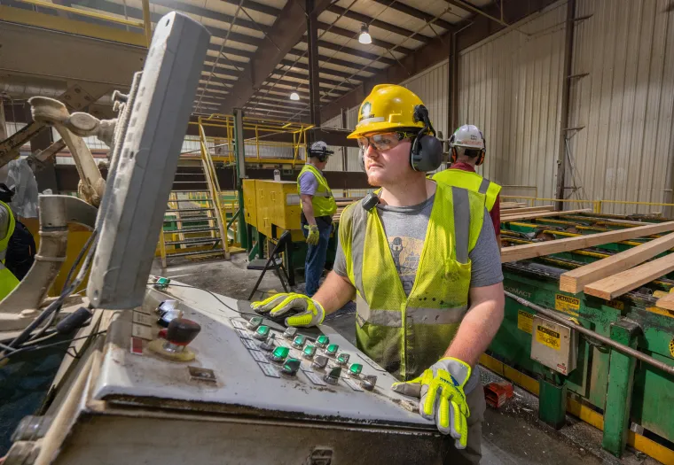 Worker in a safety helmet and high-visibility vest operates a control panel in an industrial setting, with other workers in the background.
