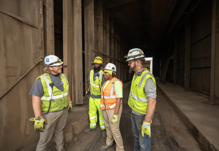 Four construction workers wearing safety gear are standing and talking inside an industrial building.