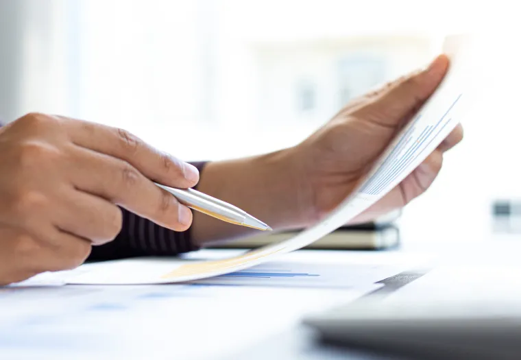 A person holding a pen in their right hand reviews a document with their left hand at a desk.