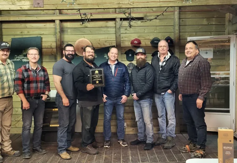 A group of eight men standing indoors, one holding a plaque. They are in a rustic wooden room with hats displayed on the wall and boxes on the floor.
