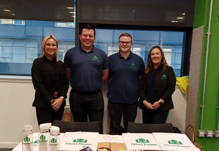 Four people stand behind a table with promotional items branded "West Fraser" in an indoor office setting.