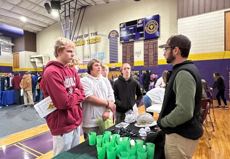A group of people engages in conversation at a table with promotional materials in a gymnasium. The table has green cups and swag bags. The gym is decorated with banners and overhead signage.