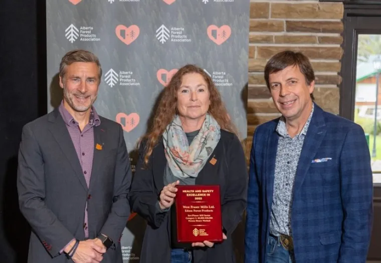 Three individuals pose together, with the middle person holding a red award plaque. They stand in front of a backdrop with logos from the Alberta Forest Products Association.