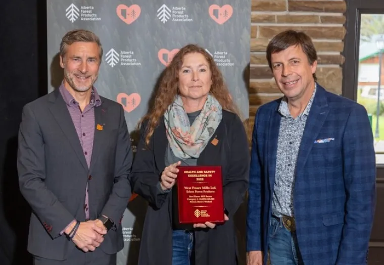 Three individuals pose together, with the middle person holding a red award plaque. They stand in front of a backdrop with logos from the Alberta Forest Products Association.