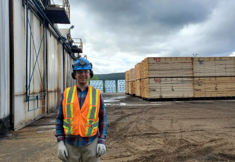A person wearing a hard hat, safety glasses, a high-visibility vest, and gloves stands in a lumber yard with stacks of wood in the background.