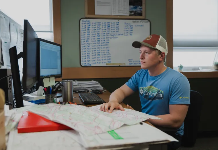 A West Fraser worker wearing a cap and a blue shirt sits at a desk with a computer, keyboard, and maps, surrounded by office materials and a whiteboard in the background.
