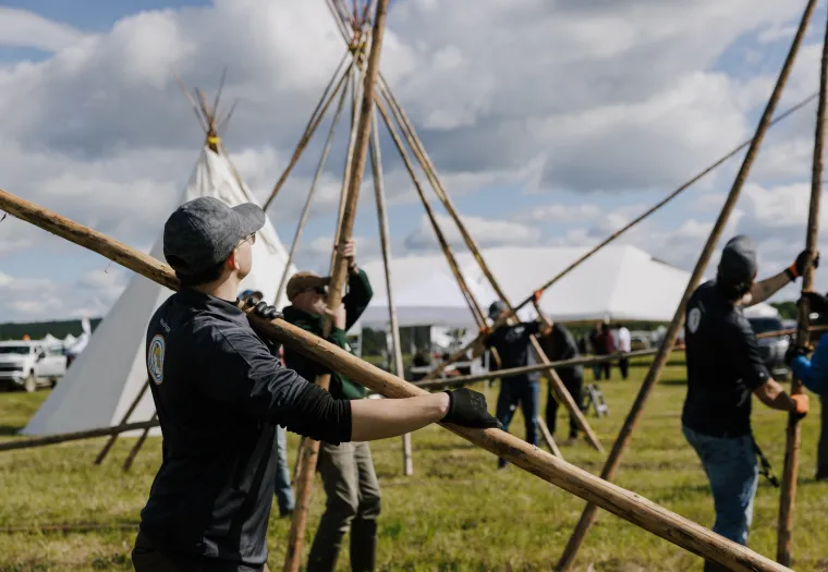 Several people work together to set up wooden poles for a large tent structure or teepee frame in an open field on a cloudy day.