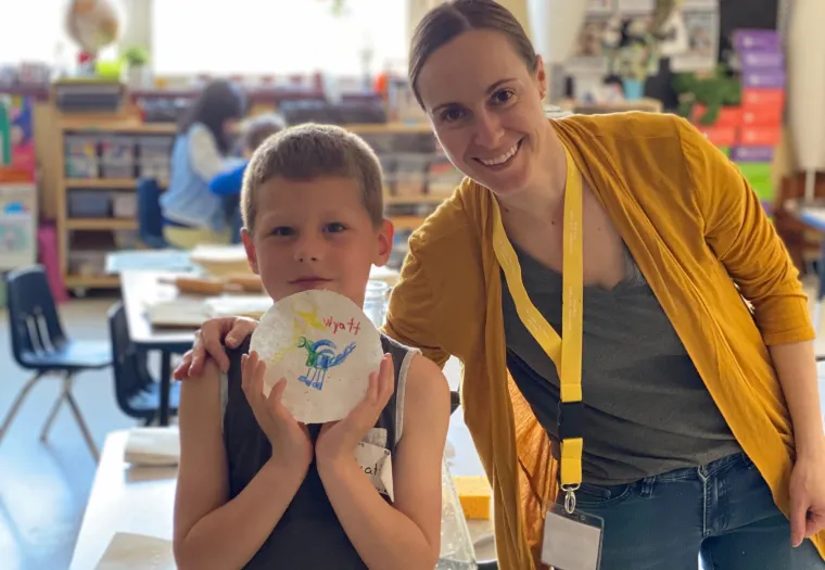A young boy holds up an art project next to a smiling woman wearing a yellow cardigan and lanyard in a classroom setting.