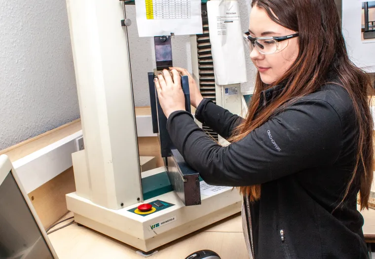 A woman wearing safety goggles operates a testing apparatus in a laboratory setting, with a computer and a caliper on the desk.