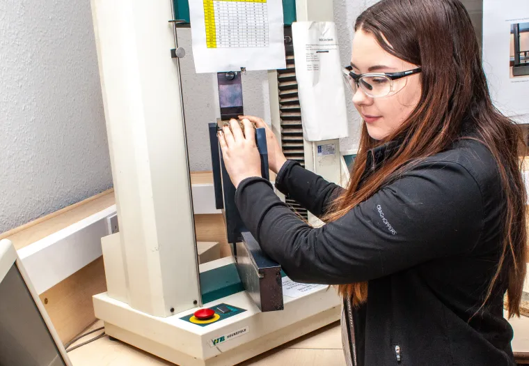 A woman wearing safety goggles operates a testing apparatus in a laboratory setting, with a computer and a caliper on the desk.
