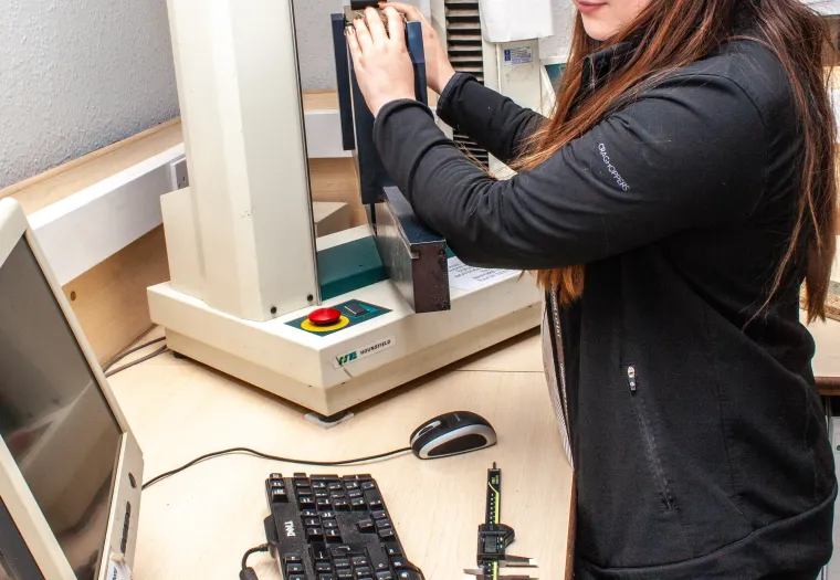 A woman wearing safety goggles operates a testing apparatus in a laboratory setting, with a computer and a caliper on the desk.