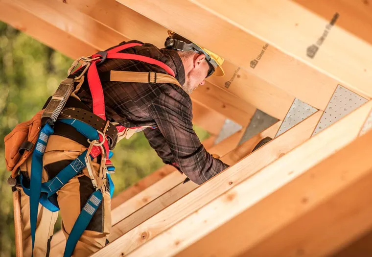 Construction worker wearing a safety harness, plaid shirt, and helmet, working on the wooden structure of a building.