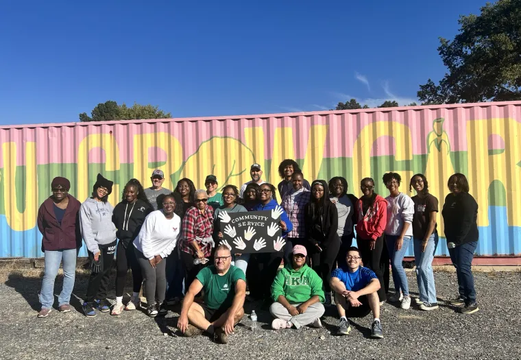 A group of people stands together in front of a colorful shipping container, holding a sign that reads "Community Service.