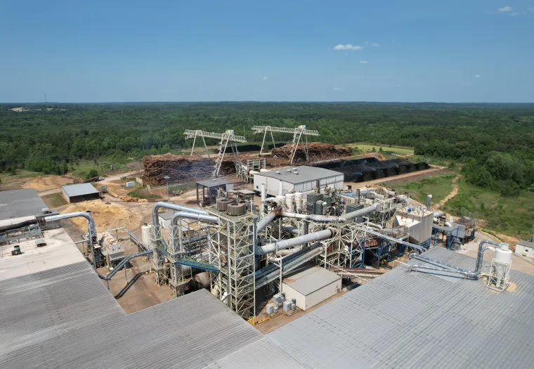 Aerial view of an industrial facility surrounded by forested land, featuring large metal structures, storage piles, and power lines under a clear blue sky.