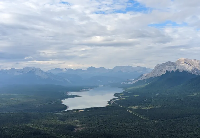 A calm lake surrounded by vast forests and distant mountain ranges under a cloudy sky.