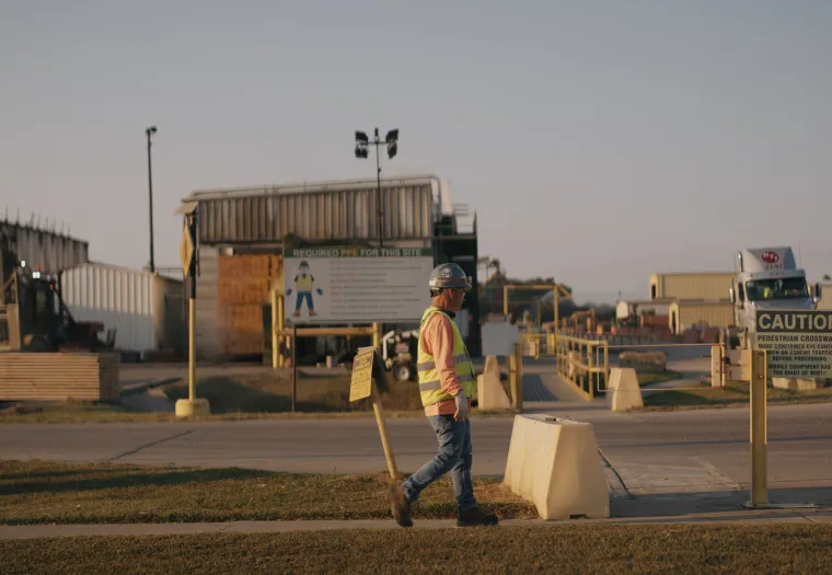 A construction worker in a high-visibility vest walks through an industrial area with caution signs and equipment in the background.